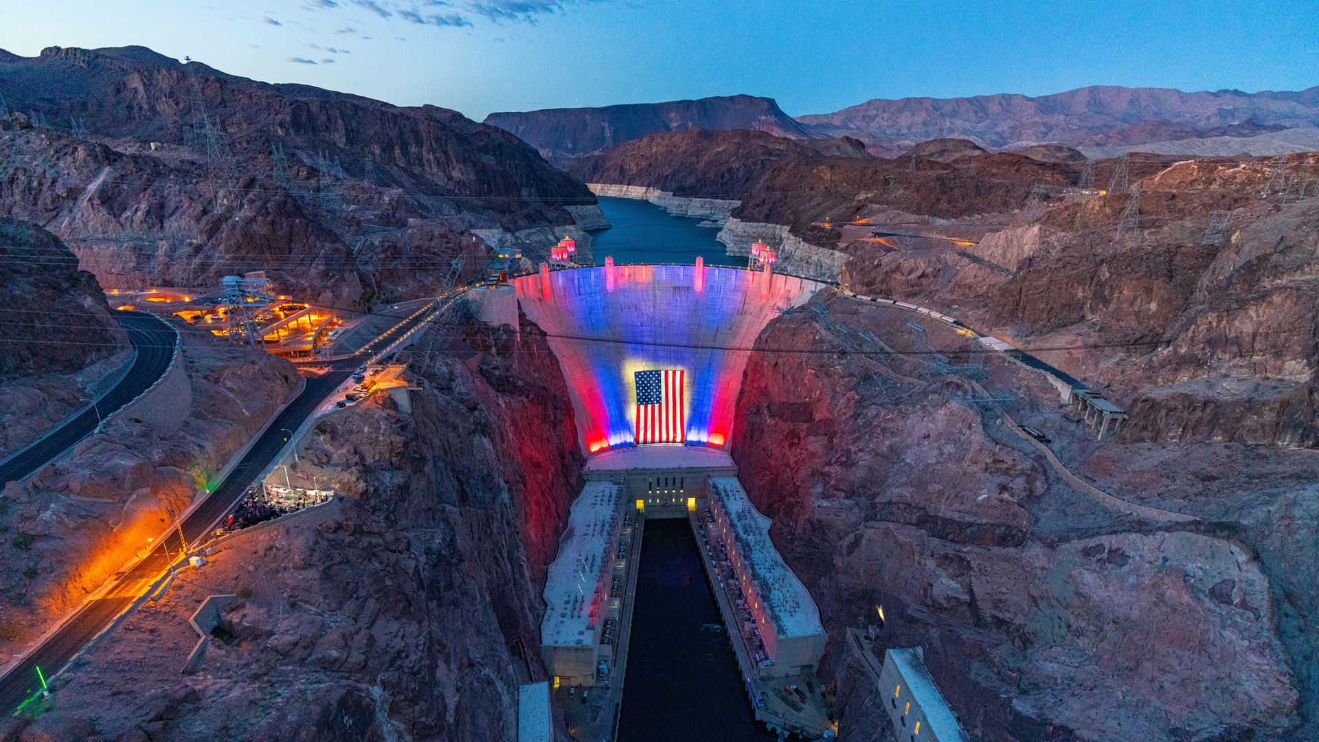 Hoover Dam illuminated in red, white, and blue with a large American flag draped across its face at dusk